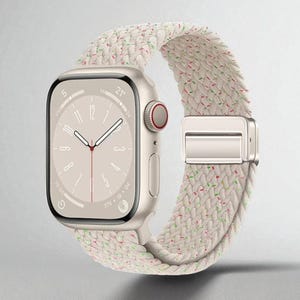 May include: A gold smartwatch with a cream-colored braided band. The watch face displays the time, with a red accent on the side button. The band has a silver clasp and is speckled with red and green threads.