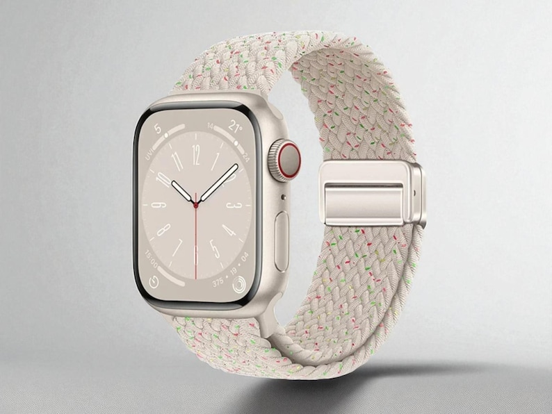 May include: A gold smartwatch with a cream-colored braided band. The watch face displays the time, with a red accent on the side button. The band has a silver clasp and is speckled with red and green threads.