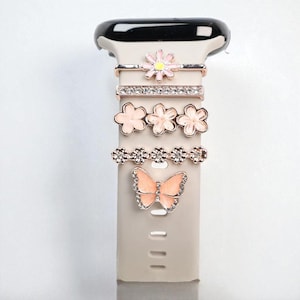 Może przedstawiać: Kolekcja ozdobnych zawieszek do paska smartwatcha. Zawieszki obejmują stokrotkę, pasek z cyrkoniami, trzy różowe kwiaty, pasek z małymi kwiatami i motyla. Zawieszki są w odcieniach różu, różowego złota i srebra.