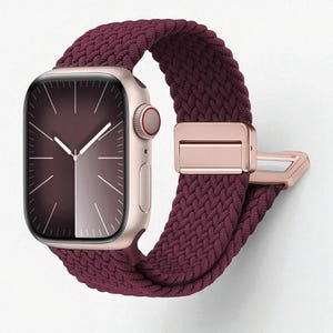 May include: A close-up of a smartwatch with a burgundy braided band and a rose gold case. The watch face displays a dark burgundy gradient with white hands and markers. The band features a rose gold clasp.