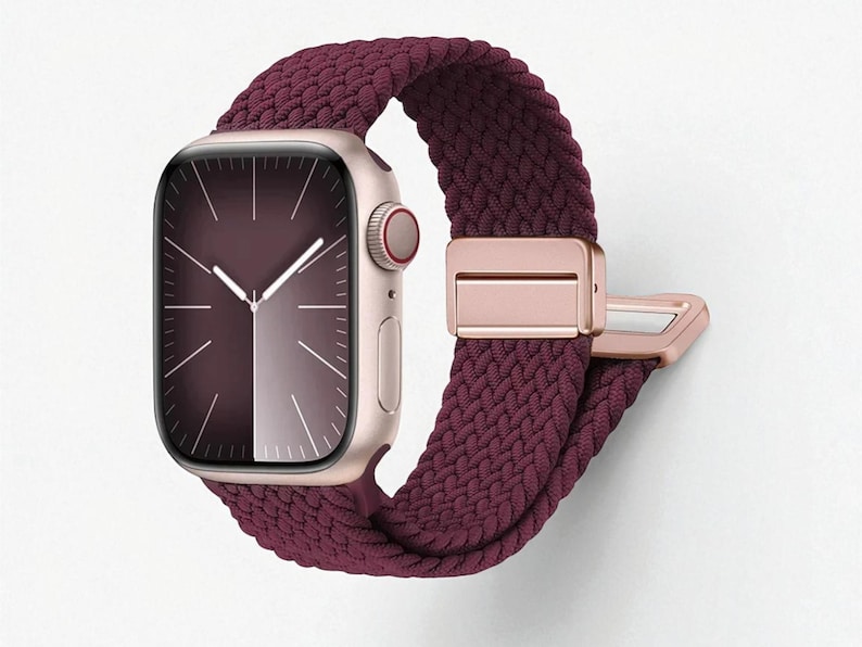 May include: A close-up of a smartwatch with a burgundy braided band and a rose gold case. The watch face displays a dark burgundy gradient with white hands and markers. The band features a rose gold clasp.