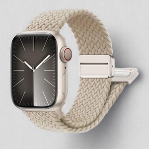 May include: A gold-colored smartwatch with a braided beige band. The watch face is divided into two tones, with a dark gray left side and a lighter gray right side. The watch has a red button and a silver clasp.
