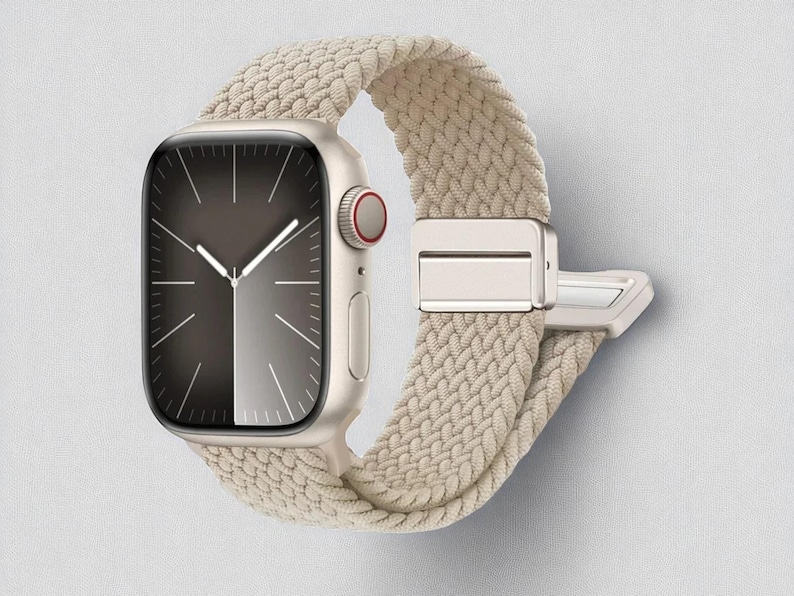 May include: A gold-colored smartwatch with a braided beige band. The watch face is divided into two tones, with a dark gray left side and a lighter gray right side. The watch has a red button and a silver clasp.