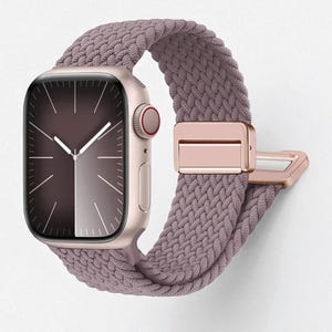 May include: A rose gold smartwatch with a braided mauve band. The watch face displays a gradient of dark and light shades. The band features a rose gold clasp. The watch is a stylish accessory.