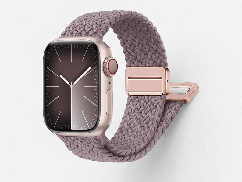 May include: A rose gold smartwatch with a braided mauve band. The watch face displays a gradient of dark and light shades. The band features a rose gold clasp. The watch is a stylish accessory.