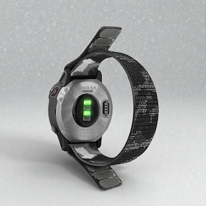 Puede incluir: Un reloj inteligente Garmin Fenix 6X negro y plateado con una correa con estampado de camuflaje. La parte inferior del reloj es visible, mostrando los sensores y el nombre del modelo. La esfera del reloj muestra luces verdes.
