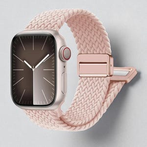 May include: A rose gold smartwatch with a braided pink band. The watch face displays a dark brown and silver gradient with hands indicating the time. The band features a matching rose gold clasp.