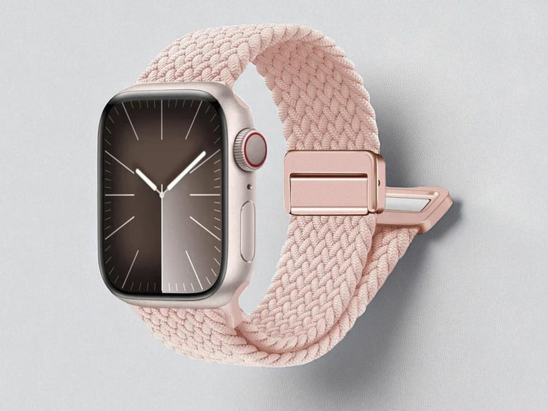 May include: A rose gold smartwatch with a braided pink band. The watch face displays a dark brown and silver gradient with hands indicating the time. The band features a matching rose gold clasp.