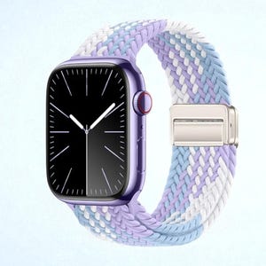 May include: A purple smartwatch with a black screen and white hands. The watch band is a braided design in shades of lavender, light blue, and white, secured with a silver clasp. The watch case has a red button.