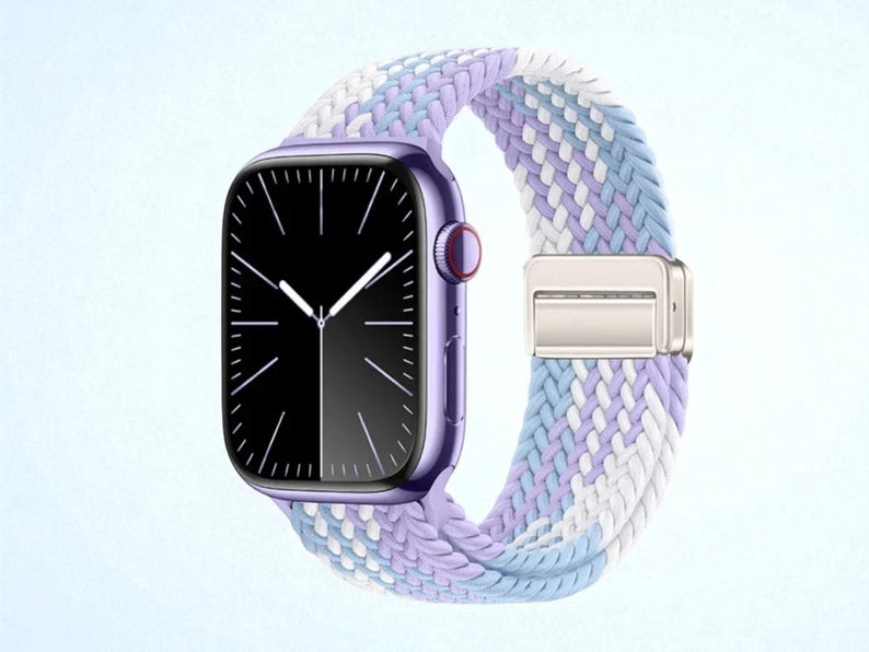 May include: A purple smartwatch with a black screen and white hands. The watch band is a braided design in shades of lavender, light blue, and white, secured with a silver clasp. The watch case has a red button.