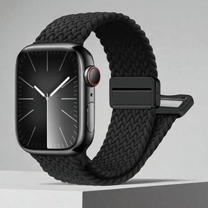 May include: A black smartwatch with a braided black band. The watch face is black with white hands and markers. The band has a secure black clasp. The watch is displayed on a white surface.