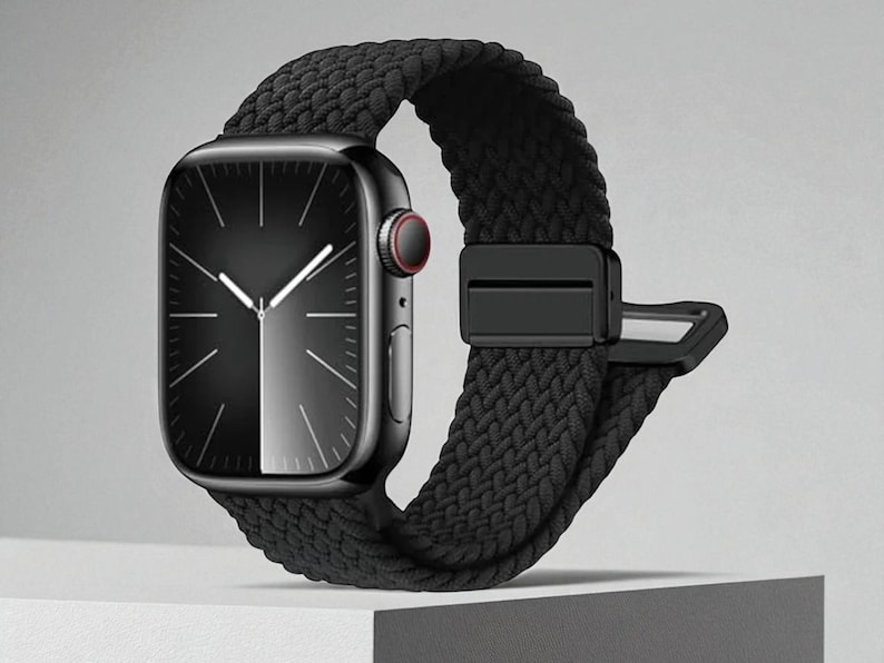 May include: A black smartwatch with a braided black band. The watch face is black with white hands and markers. The band has a secure black clasp. The watch is displayed on a white surface.