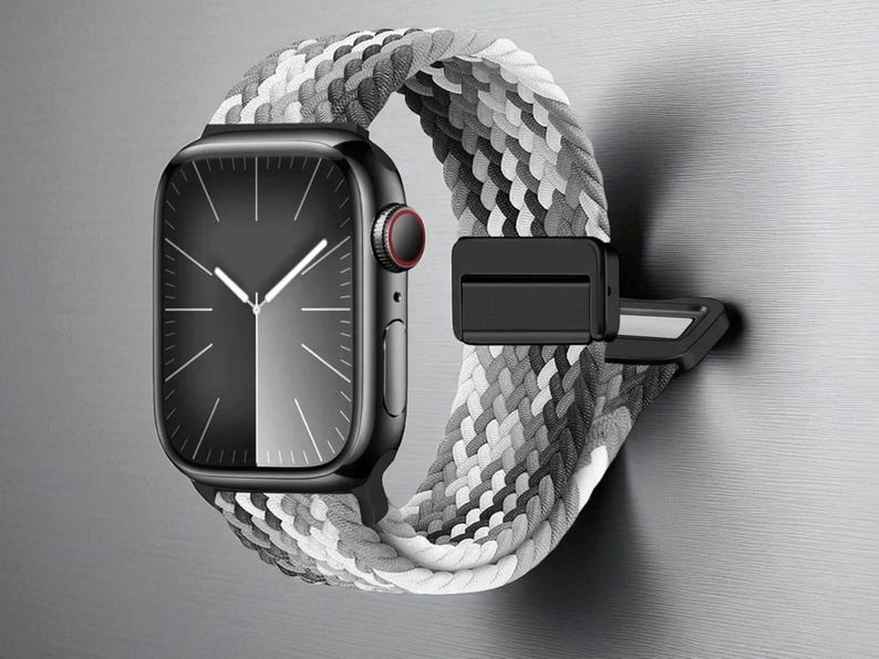 May include: A black smartwatch with a grayscale braided band. The watch face is black with white hands and markers. The band is a mix of white, gray, and black. The watch is attached to a black clasp.
