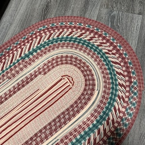 May include: Oval braided rug in shades of burgundy, cream, and teal. The rug features a plaid and striped pattern, with a braided edge. A decorative floor covering for a home.