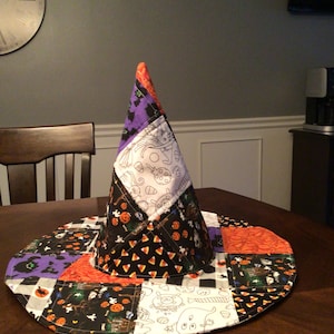 May include: A conical witch hat constructed from various quilted fabric patches featuring Halloween-themed patterns including orange pumpkins, black bats, white ghosts, and candy corn on a black, white, orange, and purple color palette.