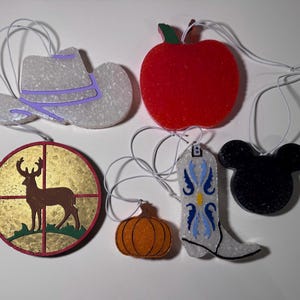 May include: A collection of six hanging ornaments featuring a white cowboy hat with purple trim, a red apple, a circular deer silhouette, an orange pumpkin, a white cowboy boot with blue details, and a black mouse-shaped silhouette.