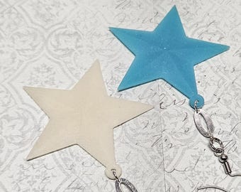 May include: Two star-shaped earrings. One is a light blue color, and the other is off-white. Each star has a silver-colored earring hook and decorative oval charm. The earrings are on a patterned background.