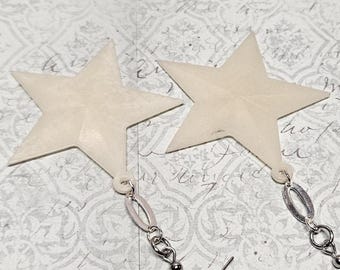 May include: A pair of cream-colored star-shaped earrings. Each star hangs from a silver-toned chain and earring hook. The earrings are set against a patterned background.