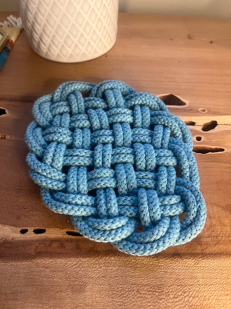 May include: A blue woven cotton coaster with a square shape and a knotted design.