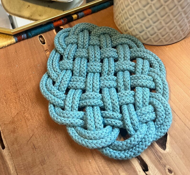 May include: A blue braided cotton trivet with a square pattern. The trivet is made of thick rope and has a woven design.