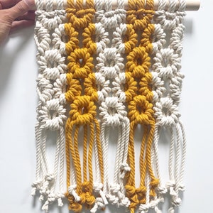 May include: A macrame wall hanging with a white and mustard yellow color scheme. The hanging features a geometric pattern with fringe at the bottom.