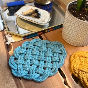 May include: Two woven rope trivets, one blue and one yellow, on a wooden surface.