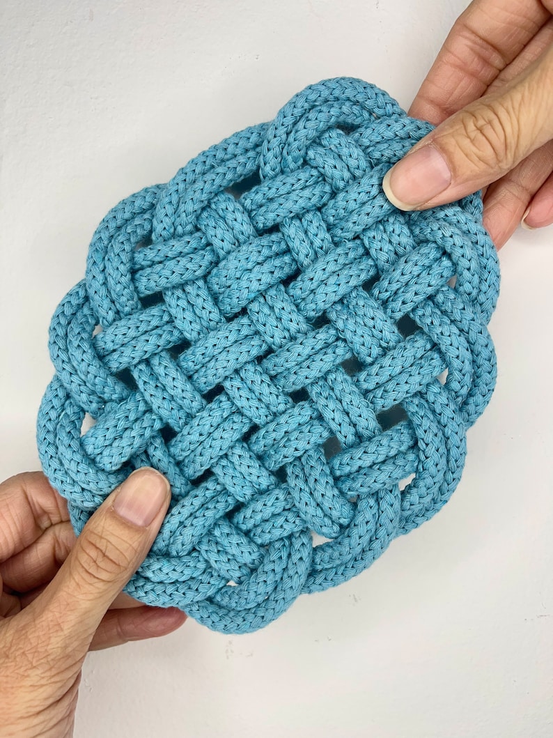 May include: A blue woven square coaster made from thick rope. The coaster is made with a knot pattern and has a slightly rounded shape.