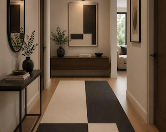 May include: A rectangular area rug featuring a geometric pattern of large black and cream-coloured blocks. The rug is positioned in a hallway with light-coloured walls, dark wood doors, a black console table, and a wall-mounted dark wood cabinet.