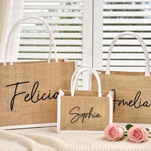 May include: Three jute tote bags with white trim and handles. The bags are personalized with the names "Felicia", "Sophia", and "Amelia" in black script. Two pink roses rest in front of the bags.