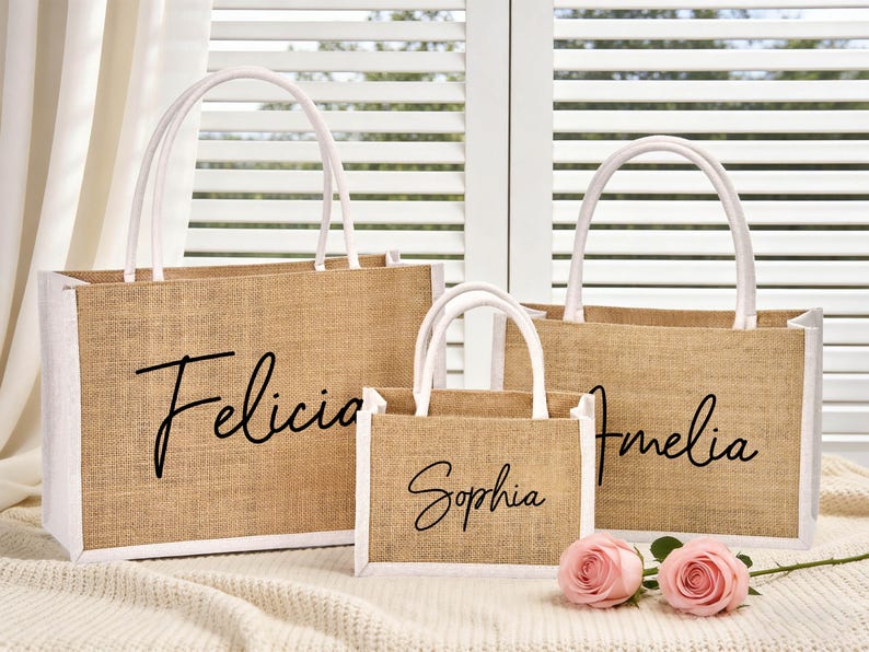 May include: Three jute tote bags with white trim and handles. The bags are personalized with the names "Felicia", "Sophia", and "Amelia" in black script. Two pink roses rest in front of the bags.