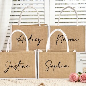 May include: Four jute tote bags with white trim and handles. Each bag has a name in elegant black script: Audrey, Naomi, Justine, and Sophia. Two pink roses rest on a cream-colored blanket.