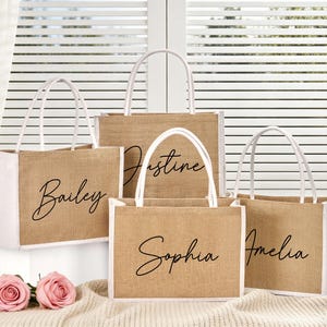 May include: Four jute tote bags with white handles and trim. Each bag has a different name in black script: Bailey, Justine, Sophia, and Amelia. The bags are on a cream-colored surface with pink roses.