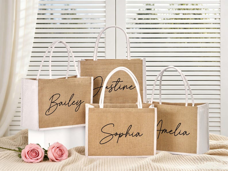 May include: Four jute tote bags with white handles and trim. Each bag has a different name in black script: Bailey, Justine, Sophia, and Amelia. The bags are on a cream-colored surface with pink roses.