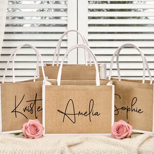 May include: A collection of jute tote bags with white accents and handles. Each bag is personalized with a name in elegant black script: Kristen, Amelia, and Sophie. Pink roses are placed in front of the bags.