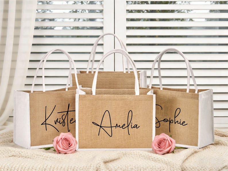 May include: A collection of jute tote bags with white accents and handles. Each bag is personalized with a name in elegant black script: Kristen, Amelia, and Sophie. Pink roses are placed in front of the bags.