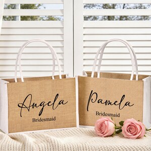 May include: Two burlap tote bags with white trim and handles. One bag says "Angela Bridesmaid" and the other says "Pamela Bridesmaid" in black script. Two pink roses are in front of the bags.