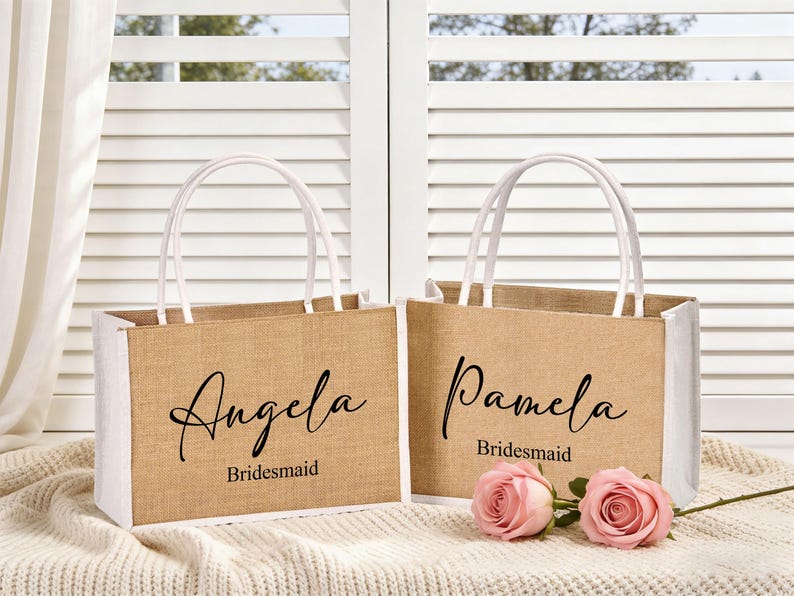 May include: Two burlap tote bags with white trim and handles. One bag says "Angela Bridesmaid" and the other says "Pamela Bridesmaid" in black script. Two pink roses are in front of the bags.