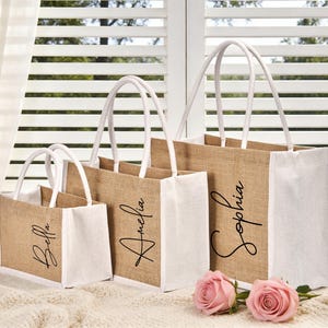May include: Three burlap tote bags with white accents and handles, each personalized with a name in elegant script: Bella, Amelia, and Sophia. Two pink roses rest in front of the bags, set against a window with white blinds.