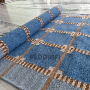 Può includere: Un tappeto blu arrotolato con un motivo geometrico di linee marroni e beige. Il tappeto è realizzato in un materiale tessuto e presenta la scritta "FLOORIFI" stampata. Il tappeto è parzialmente srotolato, rivelando la sua trama e il suo design.