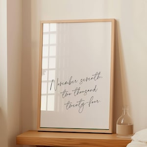 May include: A framed print with the text "November seventh, two thousand twenty-four" in elegant script. The frame is light wood, and the print is displayed on a wooden table. A small glass bottle is on the table.