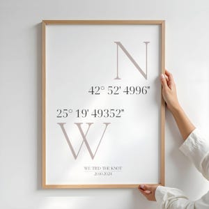 May include: A framed print with a light wood frame. The print features the letters "N" and "W" with coordinates and the text "WE TIED THE KNOT 20.10.2024" in a neutral color on a white background.