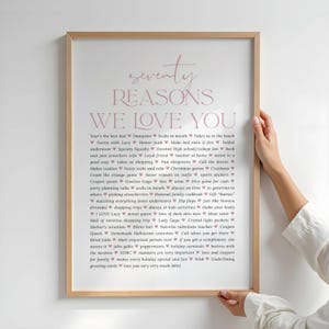 Puede incluir: Una impresión enmarcada con el texto "seventy REASONS WE LOVE YOU" en un tono rosa suave. La impresión enumera setenta razones en formato vertical, cada razón precedida por un símbolo de corazón. El marco es de color madera clara.