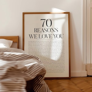 Op de afbeelding: Een ingelijste print met de tekst "70 REASONS WE LOVE YOU" in zwarte letters. De print wordt weergegeven in een slaapkamer, met een bed en gestreepte beddengoed zichtbaar op de voorgrond. De lijst is lichtbruin.
