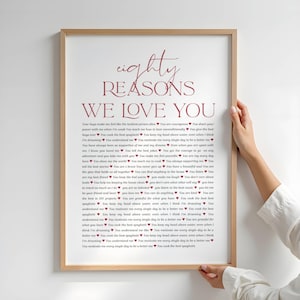 May include: A framed print with the text "Eighty Reasons We Love You" in a handwritten style. The text is in a burgundy colour and is centred on a white background. The frame is a light brown colour.