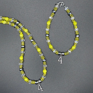 Peut inclure: Un ensemble collier et bracelet de perles jaunes, noires, transparentes et argentées. Chaque pièce comporte un charm argenté avec le chiffre 4. Les bijoux sont présentés sur un fond gris. Les perles sont mesurées en centimètres.