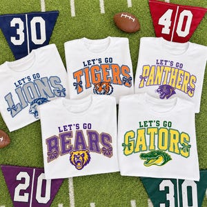 May include: Five white t-shirts featuring sports team graphics with the text Let's Go followed by team names Lions, Tigers, Panthers, Bears, and Gators. Each design includes a mascot illustration and decorative bows on the lettering.