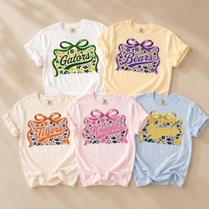May include: Five short-sleeve t-shirts in white, yellow, cream, light pink, and light blue. Each shirt features a central graphic of a ribbon bow above a floral rectangular frame containing the text Gators, Bears, Tigers, Knights, or Hornets.