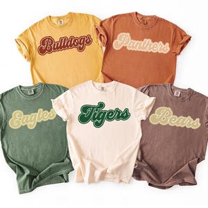Peut inclure: Cinq t-shirts à manches courtes de différentes couleurs : jaune, orange, vert, crème et marron. Chaque chemise présente un nom d'équipe dans une police rétro : "Bulldogs", "Panthers", "Eagles", "Tigers" et "Bears". Les chemises sont probablement en coton.