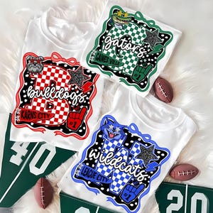 May include: White t-shirts with team logos and text, including "Bulldogs," "Gators," and "Wildcats." Each shirt features a unique design with stars, checkered patterns, and team colours. Green pennants with numbers and miniature footballs are also present.