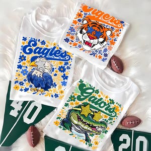 May include: White t-shirts with team graphics: "Eagles" with an eagle, "Tigers" with a tiger, and "Gators" with an alligator. Each design is surrounded by flowers. Includes green pennants with numbers and miniature footballs.